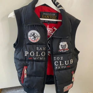 Svart dunväst från Santoz Polo Club - Snygg svart dunväst från Santoz Polo Club med broderade detaljer och dragkedja. Västen har en röd insida och flera logotyper på framsidan. Perfekt för en stilren look. Köpte den för 900kr. Priset går att diskutera😁