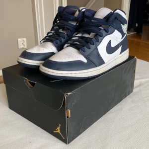 Nike Air Jordans black obsidian mids - Snygga Nike Air Jordans i en klassisk blå och vit färgkombination. Skorna har hög snörning och den ikoniska Nike-loggan på sidan. Perfekta för dig som vill ha en stilren och sportig look. Vita snören medföljer