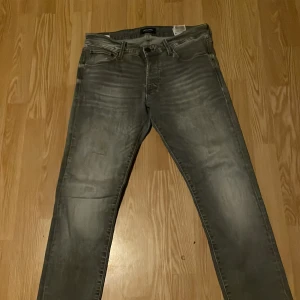 Grå jeans ifrån Jack and Jones - Snygga grå jeansbyxor med en klassisk femficksdesign och en diskret tvättad look. Byxorna har en normal passform och är perfekta för en avslappnad stil. De har en knappgylf och bälteshällor för extra funktionalitet.