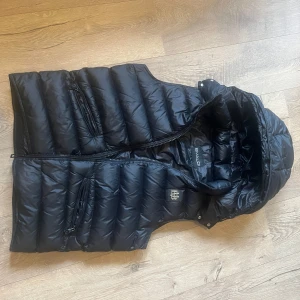 Svart dunväst från Bvaldi - Snygg svart dunväst från Bvaldi med dragkedja och flera praktiska fickor. Perfekt för kyligare dagar och ger en stilren look. Västen har en quiltad design och är idealisk för lager-på-lager-stil.