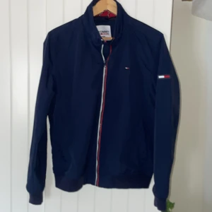 Tommy Hilfiger vårjacka, storlek m - Tommy Jeans tunn vårjacka med kraga, någorlunda vattentät, bra skydd mot vind passar även någon med storlek s. Toppskick utöver ett litet hål på insidan som syns på bild fem,  ej synligt när man har på sig jackan.