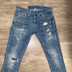 Blå jeans med slitningar - Säljer ett par blå jeans med coola slitningar och en klassisk femficksdesign. De har en knappgylf och är perfekta för en avslappnad stil. Passar bra till sneakers eller kängor.