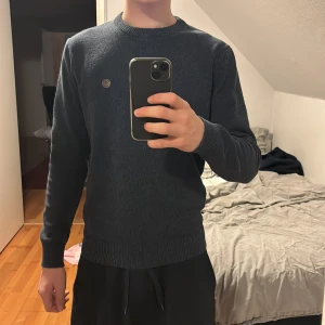 Mörkblå stickad tröja  - Säljer en stilren mörkblå stickad tröja . Tröjan har en rund halsringning och ett litet märke på bröstet. Perfekt för kyligare dagar. Hör av dig vid intresse!