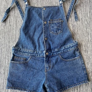 Blå jeansshorts med hängslen från OiBlue Vintage - Säljer ett par coola blå jeansshorts med hängslen från OiBlue Vintage. Shortsen har knappar framtill och flera fickor. Insidan har ett mönster med röda dödskallar. Perfekta för en avslappnad stil! Hängslejeansshorts i strl S från Oni Blue vintage denim brand.  Sparsamt använda och i fint begagnat skick.  Mått Midjan: ca 43 cm Längd från grenen och ner: ca 7 cm  97% bomull och 3% polyuretan   Kommer från ett djur och rökfritt hem.