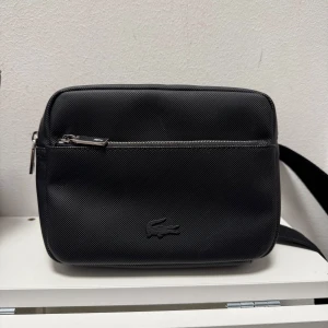 Svart axelväska från Lacoste - Snygg svart axelväska från Lacoste med justerbar rem och dragkedja. Väskan har en stilren design med det klassiska Lacoste-logotypen på framsidan. Perfekt för att bära dina dagliga nödvändigheter med stil. Pris kan diskuteras