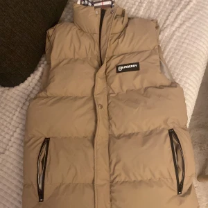Beige dunväst från Burberry - Snygg beige dunväst från Burberry med klassiskt rutigt foder. Västen har en hög krage och dragkedja framtill. Perfekt för lager-på-lager under kyliga dagar. Märkeslogga på bröstet ger en stilren touch. Storleken är M passar bra för S sitter bra. 