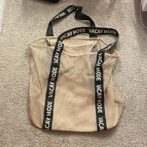 Beige nätväska med text - Säljer en beige nätväska med svarta handtag där det står 'VACAY MODE'. Perfekt för en avslappnad dag på stranden eller en snabb shoppingtur. Väskan är rymlig och lätt att bära med sig.