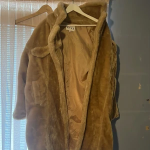 Beige pälsjacka från NA-KD - Säljer en supermjuk och lång beige pälsjacka från NA-KD. Jackan har en oversized passform och är perfekt för kyliga dagar. Den har långa ärmar och en stilren design som ger en lyxig känsla. Perfekt för att hålla sig varm och snygg under vintern!
