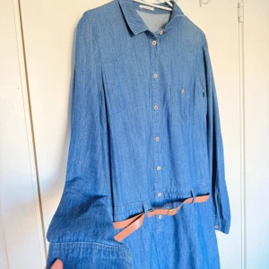 Blå jeansklänning med bälte - Använd få gånger. Snygg blå jeansklänning med långärmad design och knappar framtill. Klänningen har ett brunt bälte i midjan för en stilren look. Perfekt för en avslappnad stil med en touch av elegans.