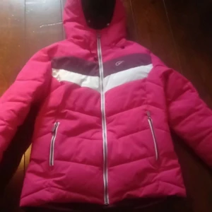 Rosa dunjacka med dragkedja - Säljer en snygg rosa vinterjacka med vit och lila detalj över bröstet. Jackan har en dragkedja framtill och två fickor med dragkedjor och en armficka. Perfekt för kyliga dagar eller en dag i skidbacken.
