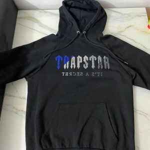 Svart hoodie från Trapstar - Säljer en svart hoodie från Trapstar med texten 'TRAPSTAR' i blått och grått på framsidan. Hoodien har en klassisk känguruficka och justerbar huva med dragsko. Perfekt för en avslappnad stil.