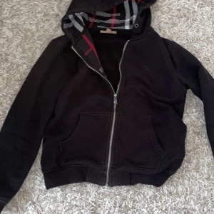 Burberry kofta - Säljer en stilren svart hoodie från Burberry då jag inte längre använder den. Dragkedjan kan vara lite trög men den funkar som den ska. Den är i storlek M men jag skulle säga den passar bättre till dig med Storlek S.