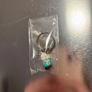 Nyckelring med kaffemugg - Säljer en unik nyckelring med en liten kaffemugg i plast. Muggen har ett lock och ett blått band runt om. Perfekt för kaffeälskare som vill ha en rolig accessoar till sina nycklar.