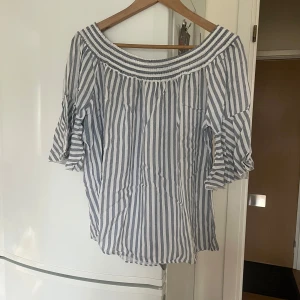 Randig offshoulder blus - Säljer en snygg randig offshoulder blus i blått och vitt. Blusen har trekvartsärmar och en luftig passform, perfekt för varma dagar. Den har en elastisk kant vid axlarna för en bekväm passform. Perfekt för en somrig look! 🌞