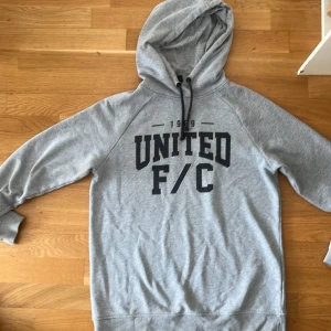 Grå hoodie med tryck - Säljer en grå hoodie med texten 'UNITED F/C' tryckt på framsidan. Tröjan har en klassisk huva med dragsko och långa ärmar. Perfekt för en avslappnad stil.