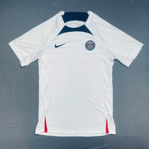 Vit PSG fotbollströja - Snygg vit PSG fotbollströja från Nike med korta ärmar och klubbens emblem på bröstet. Tröjan har mörkblåa detaljer vid halsen och röda inslag vid sidorna. Perfekt för alla fotbollsfans!