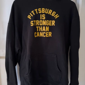 Svart hoodie från Steel City - Säljer en svart hoodie från Steel City med texten 'Pittsburgh is stronger than cancer' i gult på framsidan. 