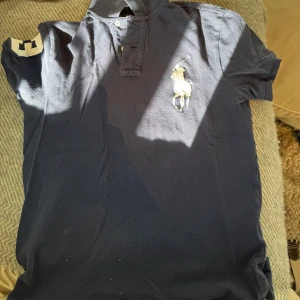 Mörkblå pikétröja från Ralph Lauren - Säljer en klassisk mörkblå pikétröja från Ralph Lauren med en broderad logga på bröstet. Tröjan har korta ärmar och en knappslå framtill. Perfekt för en stilren look.