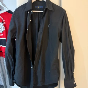 Svart skjorta från Ralph Lauren - Säljer en stilren svart skjorta från Ralph Lauren i storlek L men passar även M. Skjortan har en slim fit passform och är dekorerad med en liten broderad logotyp på bröstet. Perfekt för en elegant look.