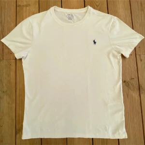 Vit t-shirt från Ralph Lauren - Säljer en klassisk vit t-shirt från Ralph Lauren med en liten broderad logga i blått på bröstet. T-shirten har en rund hals och korta ärmar, perfekt för en stilren look.