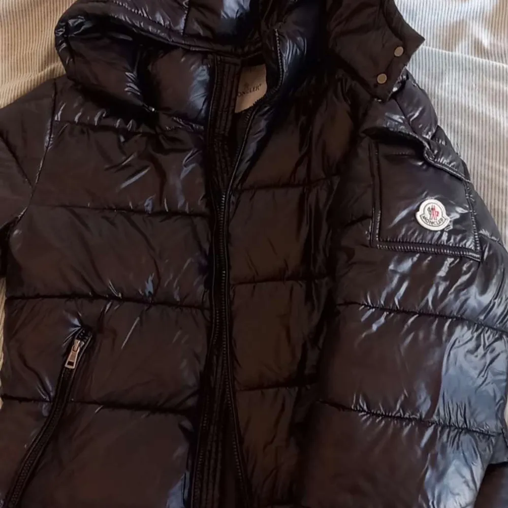 Säljer en snygg svart pufferjacka från Moncler med glansig finish. Jackan har en praktisk huva och dragkedja framtill. Perfekt för kyligare dagar med sin quiltade design och ikoniska Moncler-logga på ärmen. Logan skannar som vanligt.. Takit.