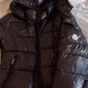 Svart pufferjacka från Moncler - Säljer en snygg svart pufferjacka från Moncler med glansig finish. Jackan har en praktisk huva och dragkedja framtill. Perfekt för kyligare dagar med sin quiltade design och ikoniska Moncler-logga på ärmen. Logan skannar som vanligt.