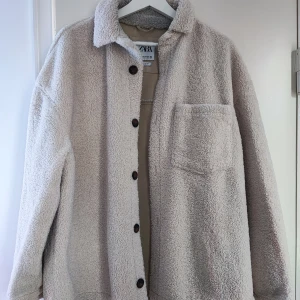 Beige overshirt i teddy från Zara - Mysig beige teddy overshirt från Zara med knappar framtill och en bröstficka. Perfekt för kyliga dagar med sin mjuka och fluffiga textur. Overshirten har en klassisk krage och långa ärmar.