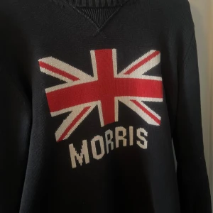 Svart tröja från Morris med Union Jack - Säljer en svart tröja från Morris med ett stort Union Jack-motiv och texten 'Morris' på framsidan. Tröjan har långa ärmar och ribbade detaljer vid hals och ärmslut. Perfekt för en stilren look. Priset kan diskuteras