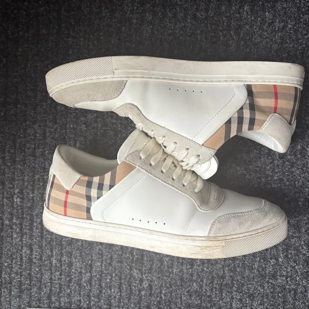 Snygga vita sneakers från Burberry med klassiskt rutigt mönster på hälen. Skorna har snörning och en stilren design med detaljer i mocka och syntetmaterial. Perfekta för en trendig look. Köpte för 6800 Allt Og ingår. Kengät.