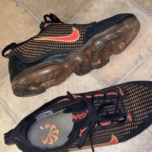 Nike Vapormax sneakers i svart och orange - Snygga Nike Vapormax sneakers i svart med orange detaljer. Skorna har en unik sula med synliga luftkuddar för extra komfort och stil. Perfekta för både träning och vardag.