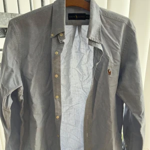Ljusblå skjorta från Ralph Lauren - Säljer en stilren grå skjorta från Ralph Lauren med klassisk krage och knappar framtill. Skjortan har en broderad logotyp på bröstet och långa ärmar. Perfekt för en elegant look.