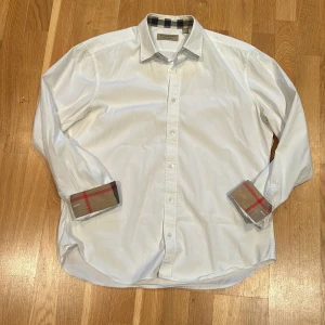 Vit skjorta från Burberry - Säljer en stilren vit skjorta från Burberry med klassiskt rutigt mönster på insidan av kragen och ärmsluten.✅ Skjortan har långa ärmar och knappar framtill. Perfekt för en elegant look. Väldigt fin skjorta med en liten fläck som inte syns om man planerar att ha ärmarna uppvikta😊Skriv till mig vid frågor och funderingar💯