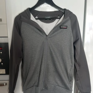 Grå huvtröja från McKenzie - Säljer en grå huvtröja från McKenzie med dragkedja och huva. Tröjan har långa ärmar och en stilren design med vita detaljer på ärmarna. Perfekt för en avslappnad stil. Pris kan diskuteras 👍