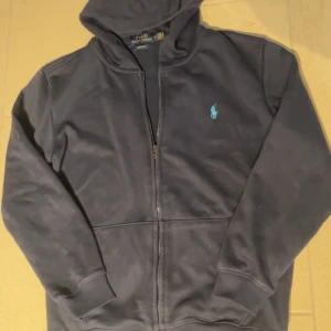 Mörkblå hoodie från Ralph Lauren - Säljer en stilren mörkblå hoodie från Ralph Lauren med dragkedja och det klassiska logotypbroderiet på bröstet. Tröjan har långa ärmar och en bekväm passform. Perfekt för en avslappnad look.