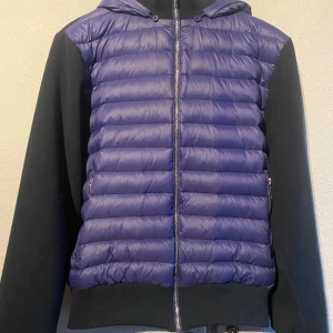 Blå och svart cardigan från Levon - Snygg cardigan från Levon i blått och svart. Jackan har en quiltad framsida med dragkedja och huva. Perfekt för kyligare dagar och perfekt för våren med sin stilrena design och bekväma passform. Storleken är S men passar även M, ny pris 1000kr, mitt pris 899kr