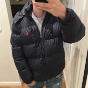 Mörkblå dunjacka från Ralph Lauren - Säljer en snygg mörkblå dunjacka från Ralph Lauren med dragkedja och ikonisk röd logga på bröstet. Jackan har långa ärmar och är perfekt för kyligare väder. Den är både stilren och funktionell. Passar 183-190
