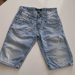 Ljusblå jeansshorts från Smog - Säljer ett par ljusblå jeansshorts från Smog. De har en klassisk femficksdesign och är perfekta för sommardagar. Shortsen har en knappgylf och bälteshällor för extra stil och funktionalitet.