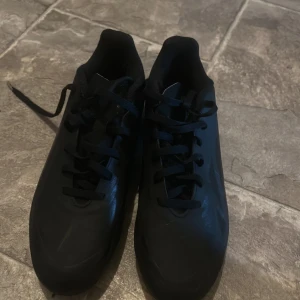 Svarta fotbollsskor från Adidas - Säljer ett par svarta fotbollsskor från Adidas med snörning och dobbar för optimalt grepp på planen. Perfekta för fotbollsträning och matcher. Skorna har en stilren design och är gjorda i syntetmaterial.