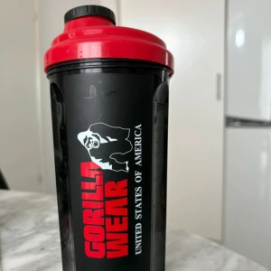 Svart shaker från Gorilla Wear - Säljer en svart shaker från Gorilla Wear med röd skruvlock och vit logotyp. Perfekt för proteinshakes och smoothies. Inkluderar ett vitt nät för bättre blandning. Lätt att rengöra och praktisk för gymmet eller på språng. Aldrig använd