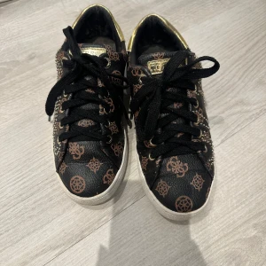 Svarta sneakers med mönster från Guess - Snygga svarta sneakers från Guess med brunt mönster och gulddetaljer. Skorna har snörning och är dekorerade med små pärlor. Perfekta för att ge din outfit en unik touch.