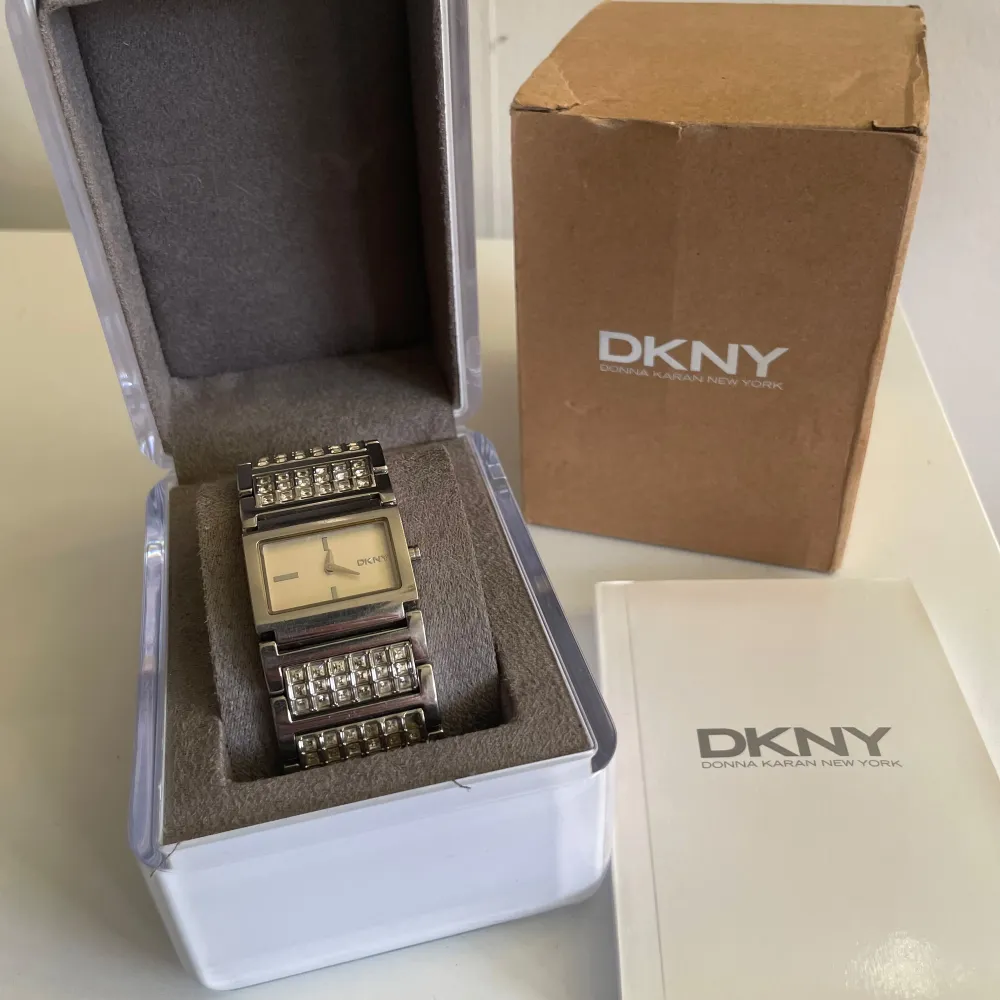 Snygg och elegant klocka från DKNY i silverfärg. Klockan har en rektangulär urtavla och ett armband dekorerat med glittriga detaljer. Obs! Batteriet måste bytas! Perfekt för att ge en touch av glamour till din stil.. Asusteet.