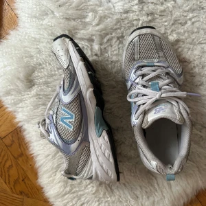 Grå och blå sneakers från New Balance - Säljer ett par grå och blå sneakers från New Balance med snörning. Skorna har en sportig design med meshdetaljer för extra andningsförmåga. Perfekta för en aktiv livsstil.