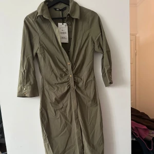 Olivgrön klänning från Zara - Snygg olivgrön klänning från Zara med trekvartsärmar och knappar framtill. Klänningen har en elegant rynkdetalj i midjan som ger en smickrande passform. Perfekt för en stilren look.
