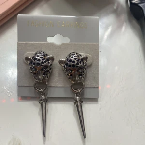 Silvriga leopardörhängen med spetsdetalj - Snygga örhängen i form av leopardhuvuden i silver med svarta fläckar. De har en hängande spetsdetalj som ger en edgy look. Perfekta för att ge din outfit en unik touch.väldigt populära nu i 2025.Hör gärna av dig vid minsta intresse.