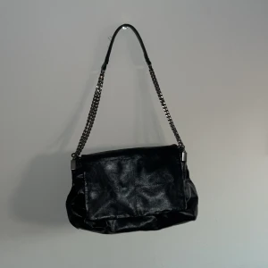 Svart axelväska med kedjedetaljer från Zara - Snygg svart axelväska i skinn med kedjedetaljer på axelremmen. Väskan har flera fack med dragkedja och en praktisk magnetknapp för enkel stängning. Perfekt för att hålla ordning på dina saker med stil.