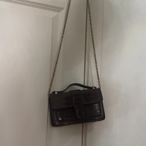 Brun axelväska med krokodilmönster - Snygg brun axelväska i skinn med krokodilmönster och guldfärgade detaljer. Väskan har en kedjerem och ett praktiskt spänne framtill. Perfekt för att ge en elegant touch till din outfit.