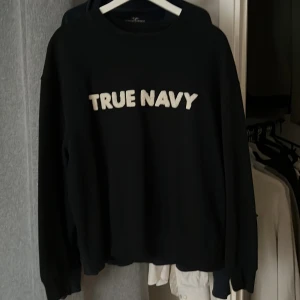 Mörkblå tröja med text från East West - Säljer en mörkblå tröja från East West med texten 'TRUE NAVY' i vitt på framsidan. Tröjan e lite kort för mig och jag är 181 cm lång. Perfekt för en lite mer avslappnad stil. 