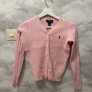  kofta från Ralph Lauren - Säljer en söt rosa kabelstickad kofta från Ralph Lauren. Den har knappar framtill och det klassiska logotypbroderiet på bröstet. Perfekt för en stilren och mysig look. 💖storlek L i barnstorlek!!! så egentligen xs/s💗kolla upp deras storlekar.