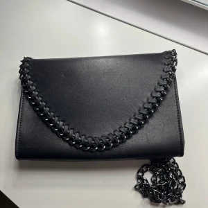 Svart väska från Tiamo - Snygg svart kuvertväska från Tiamo med kedjedetaljer längs kanten. Väskan har en praktisk dragkedja inuti för säker förvaring. Perfekt för en stilren look.
