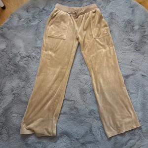 Beige mjukisbyxor från Juicy Couture - Junior 8-9 år Snygga beige mjukisbyxor från Juicy Couture med bred resår och knytband i midjan. Byxorna har praktiska fickor fram och bak med knappdetaljer. Perfekta för en avslappnad stil.